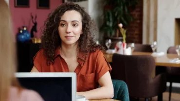 Woman with curly hair in red blouse talking at a cafe.