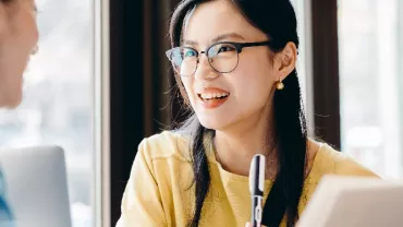 Two women having a conversation at a table, one holding a pen and smiling.