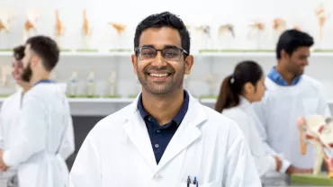 A smiling lab technician in a white coat stands in a science classroom with colleagues working in the background.