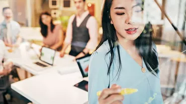 Woman writing on glass board in office meeting with colleagues watching.