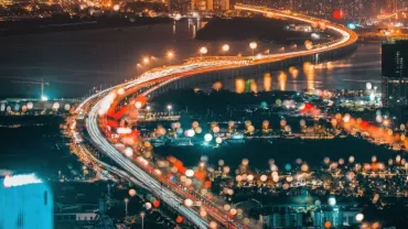 Aerial view of a city at night with a brightly lit bridge and blurred lights in the foreground.