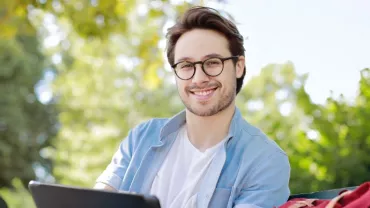 Man with glasses smiling while sitting outside with greenery in the background.