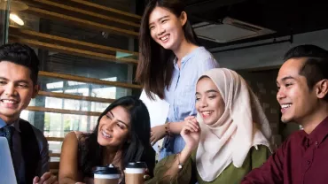 Group of five people smiling and looking at a laptop screen in an office setting.