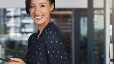 Smiling woman in a black blouse holding a smartphone indoors.