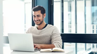 Man smiling while using a laptop at a desk with a notebook open.