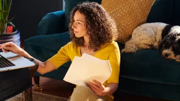 Woman working with a laptop and papers, dog resting on couch in sunlit room.