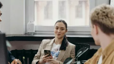 Three people having a meeting in an office, two are sitting and one is holding a phone.