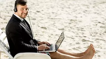 Man in a suit using a laptop on a beach chair with headphones.